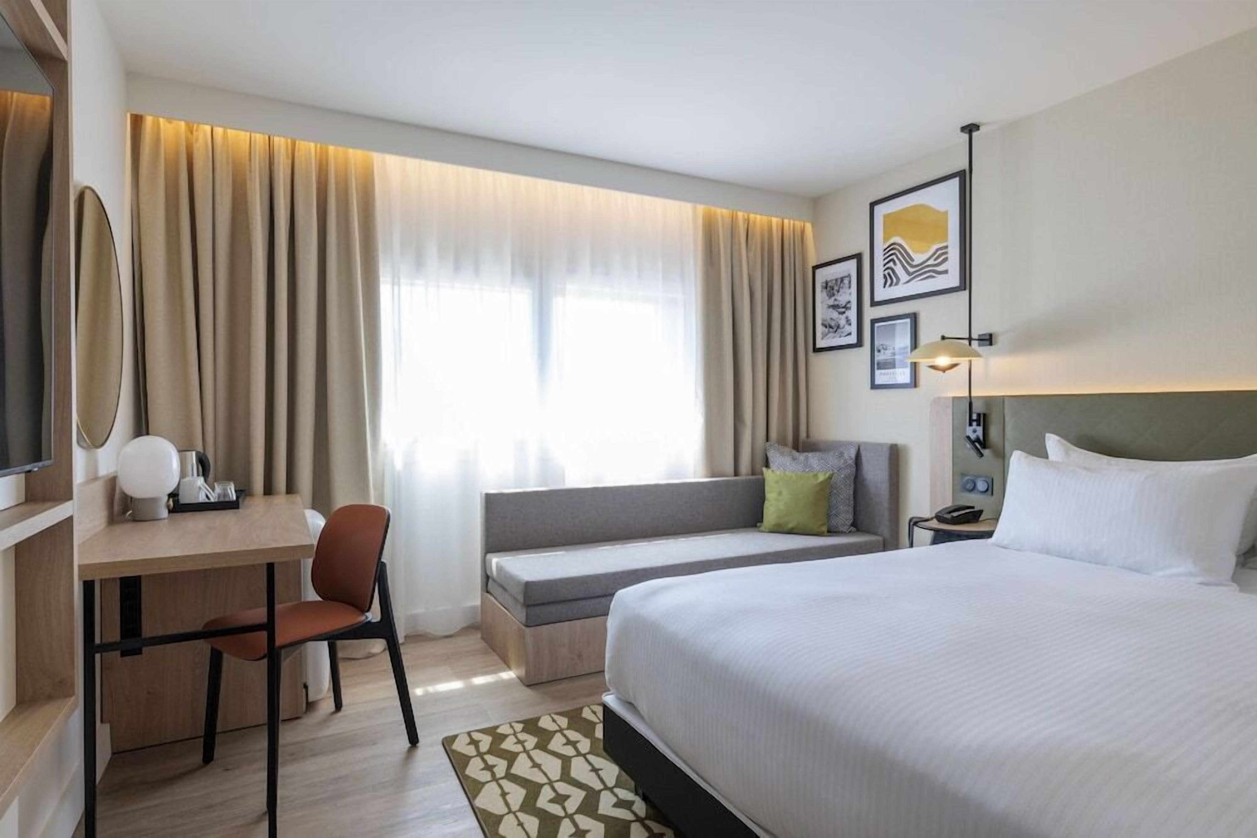 Une chambre d’hôtel avec un lit, un bureau, une chaise, un canapé et une fenêtre avec des rideaux.