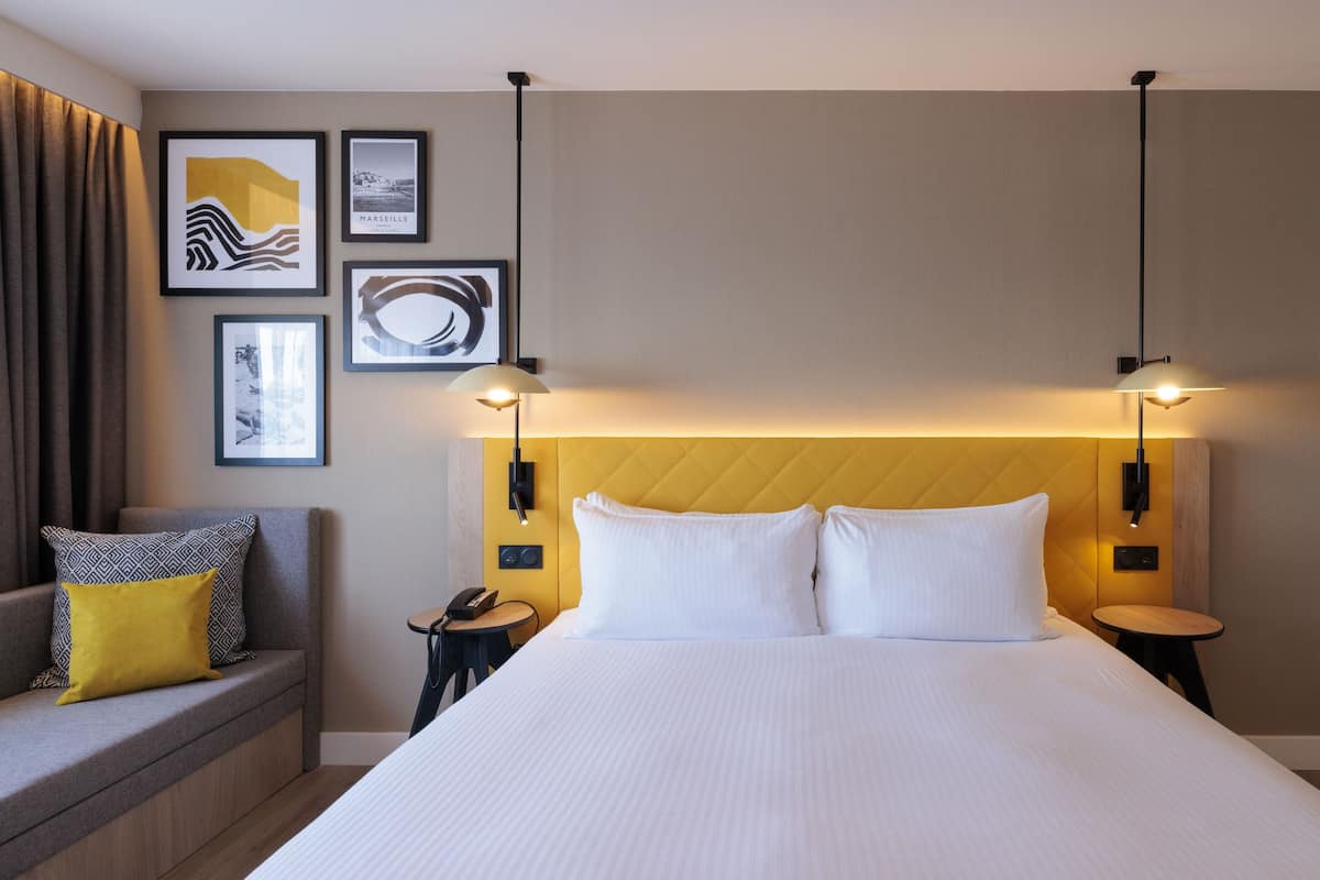 A hotel room with a large bed, a sofa with cushions, a small table, and wall-mounted artwork.