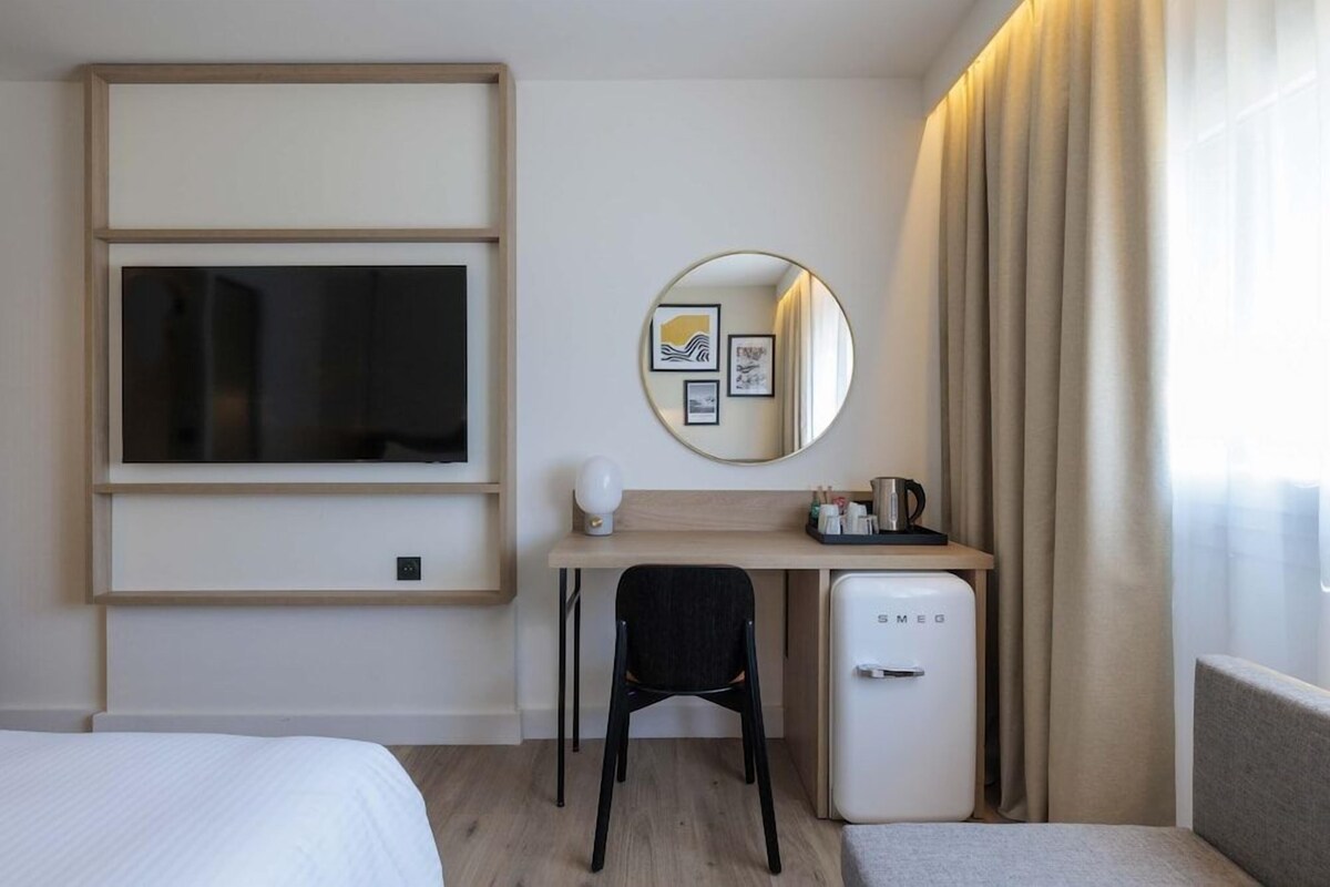 Habitación de hotel con cama, escritorio con silla, refrigerador pequeño, espejo redondo y ventana con cortinas.