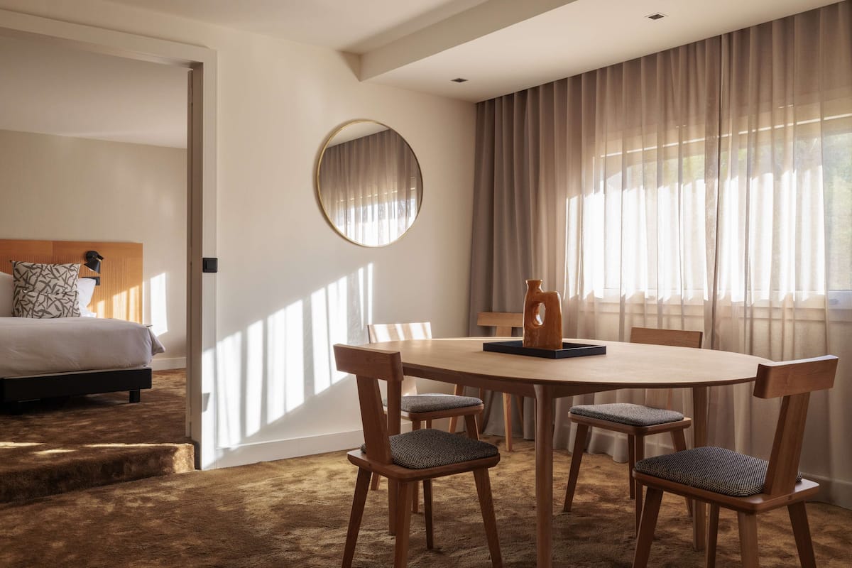 A hotel room with a bed, a round mirror, a dining table with chairs, and a view of the outdoors through large windows.