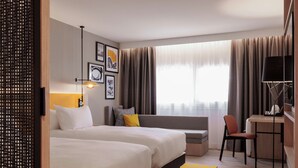 Quarto de hotel com duas camas, uma escrivaninha, uma cadeira, um sofá e uma janela grande com cortinas.