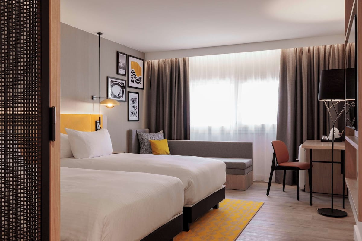A hotel room with two beds, a desk, a chair, a sofa, and a large window with curtains.