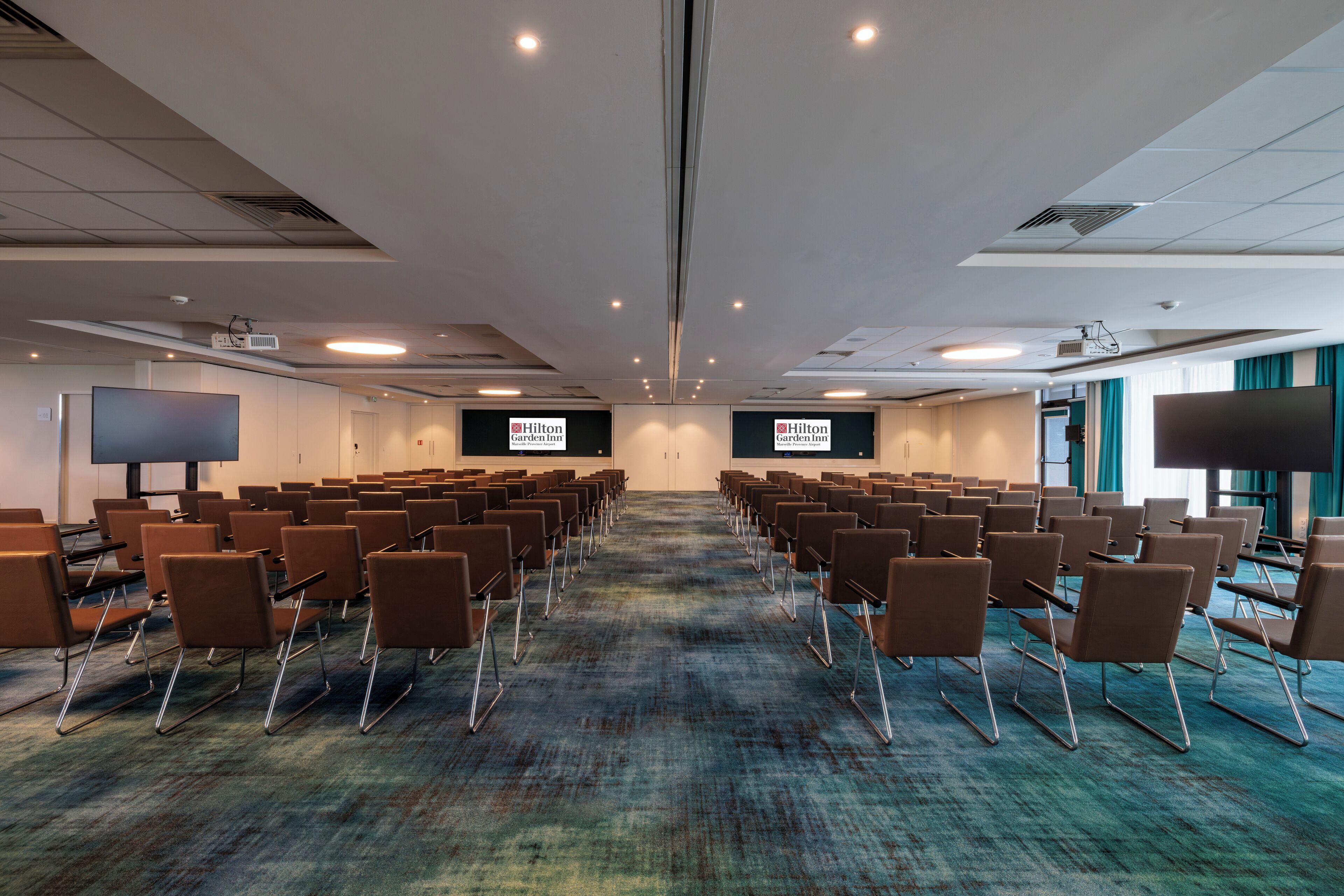 Un salón de conferencias con filas de sillas, piso alfombrado y dos pantallas mostrando el logotipo de Hilton Garden Inn.
