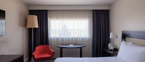 Quarto de hotel com cama, uma cadeira vermelha, uma mesa redonda pequena, mesa de cabeceira com abajur e janela com cortinas.