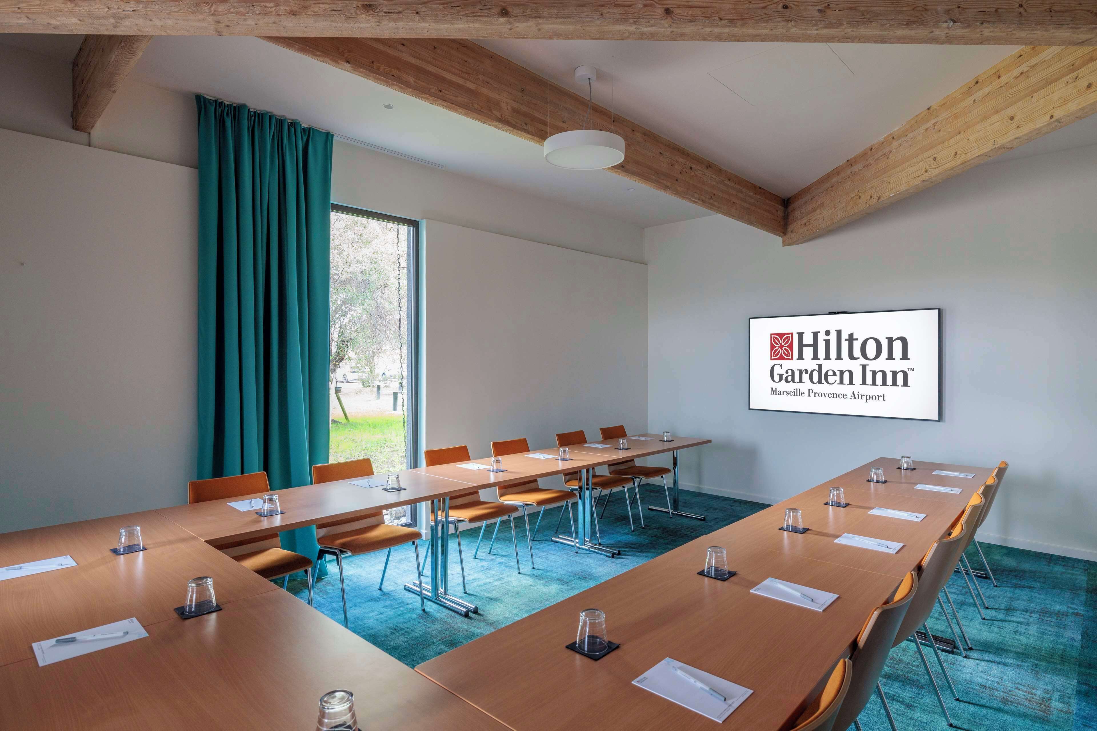 A conference room with a long table, chairs, and a large window with curtains. A sign for Hilton Garden Inn is visible on the wall.