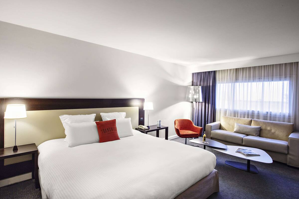 A hotel room with a large bed, a sofa, a coffee table, a chair, and a lamp.