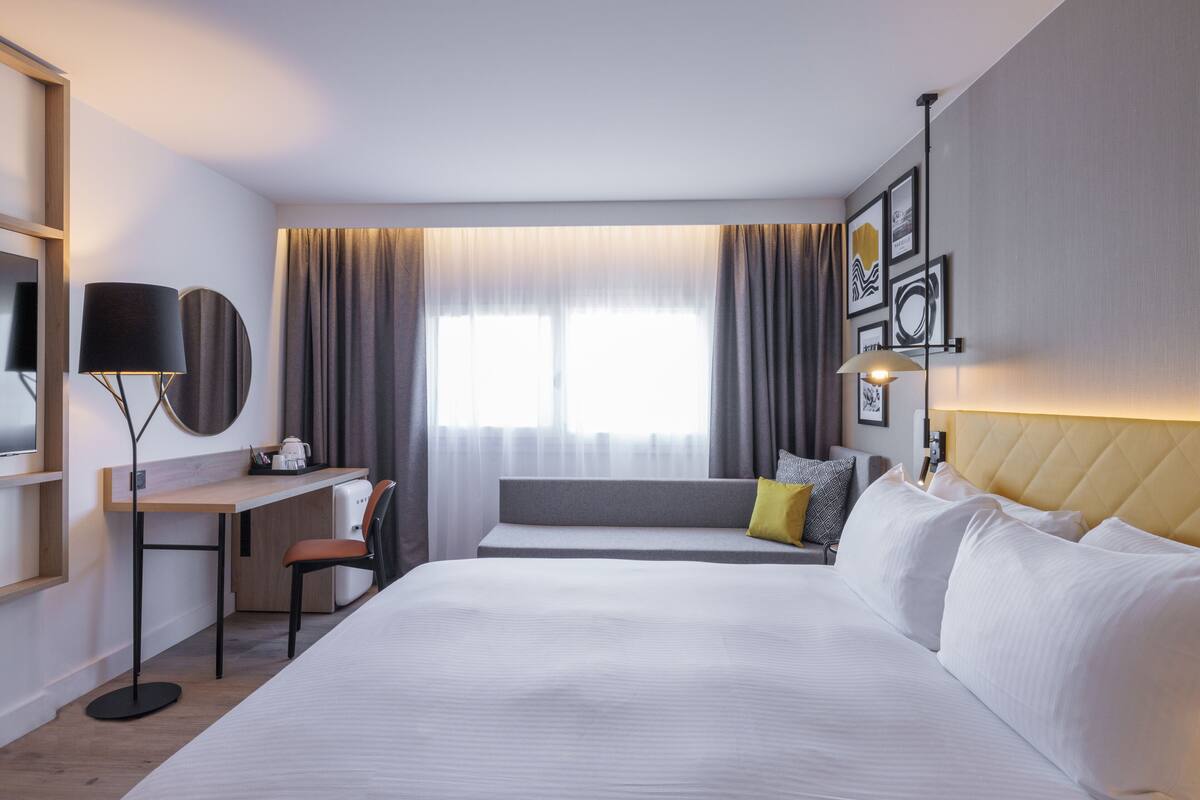 A hotel room with a large bed, a desk with a chair, a TV, and a window with curtains.
