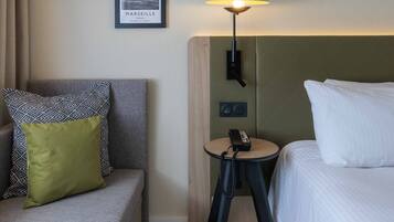 A hotel room with a bed, a sofa, a small table with a lamp, and three framed pictures on the wall.