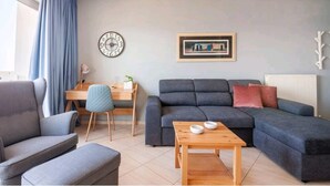 A modern living room with a grey sofa, a wooden coffee table, a desk with a chair, and a wall clock. - Valeria Sea View Apartment in Rethymno, Crete (Rethymno)