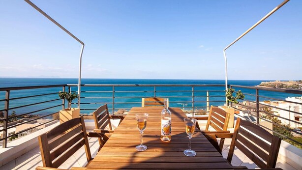 A wooden table with two wine glasses and a bottle, set against a backdrop of the sea and a clear sky. - Valeria Sea View Apartment in Rethymno, Crete (Rethymno)