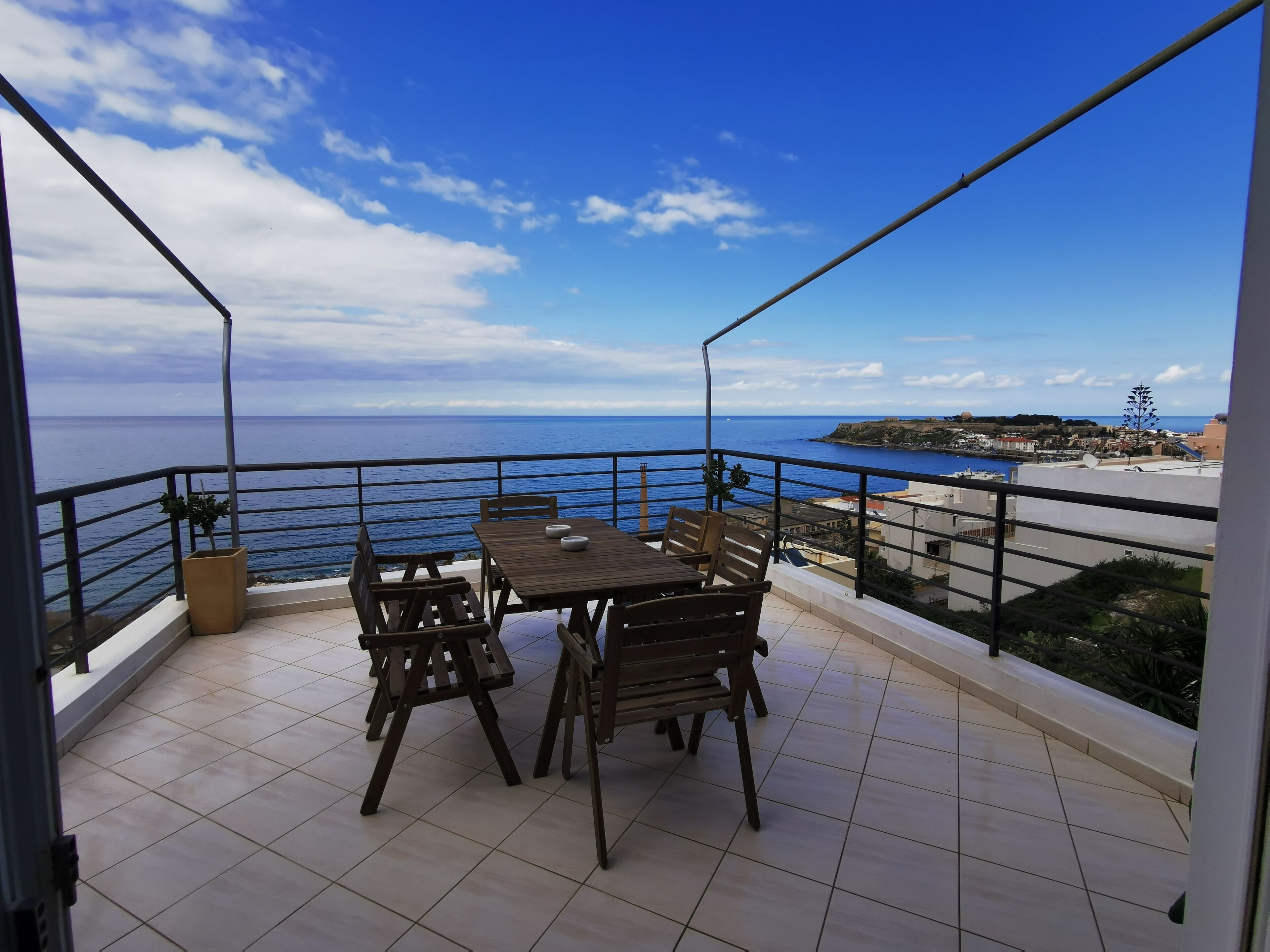 A balcony with a wooden table and chairs, offering a view of the sea and a distant coastline.