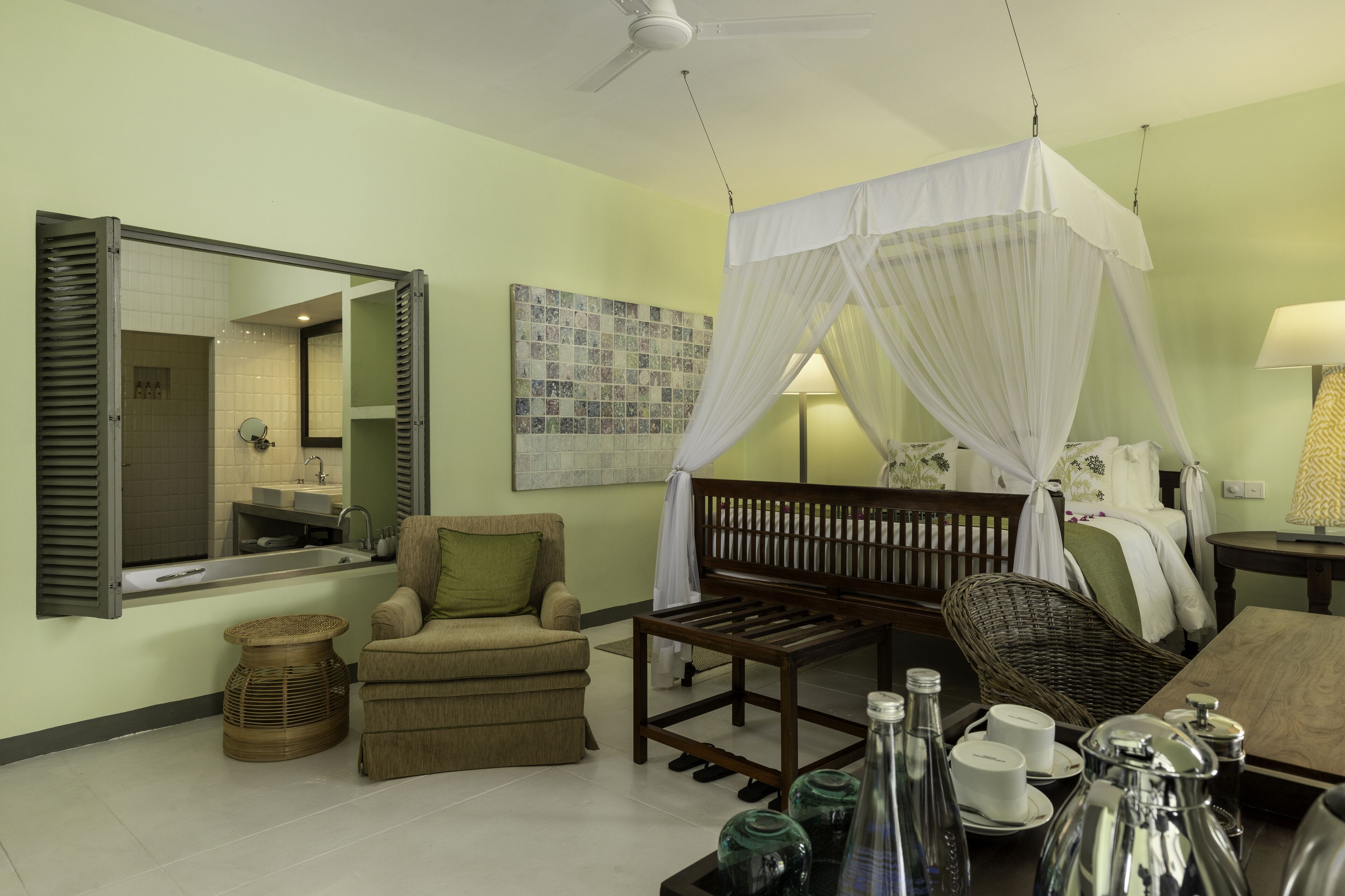 A room with a bed, a chair, a small table with items, a mirror, a wall with a picture, and a bathroom with a sink and mirror.
