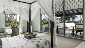 A bedroom with a canopy bed, a wicker chair, a small table with a lamp, and a view of the outdoors through large windows. - The Villa Bentota by KK Collection (Bentota)