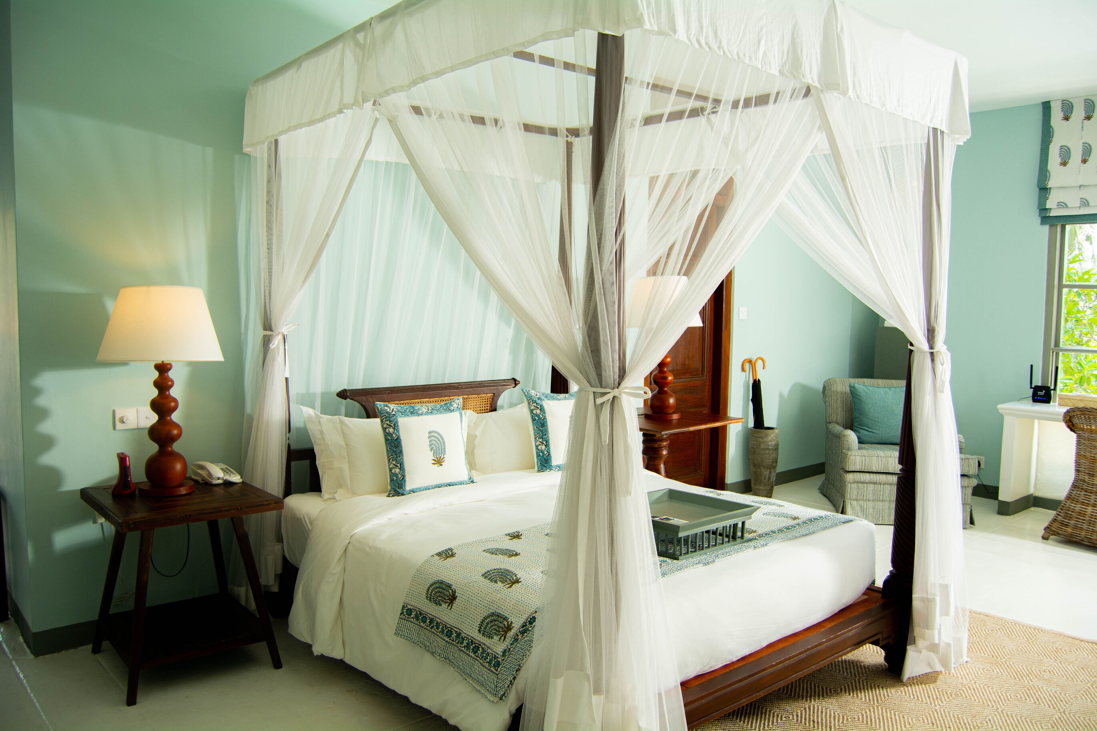Una cama con dosel y cortinas, con colcha blanca y detalles azules, una mesita de noche con lámpara y una silla de mimbre en una habitación con paredes azul claro.