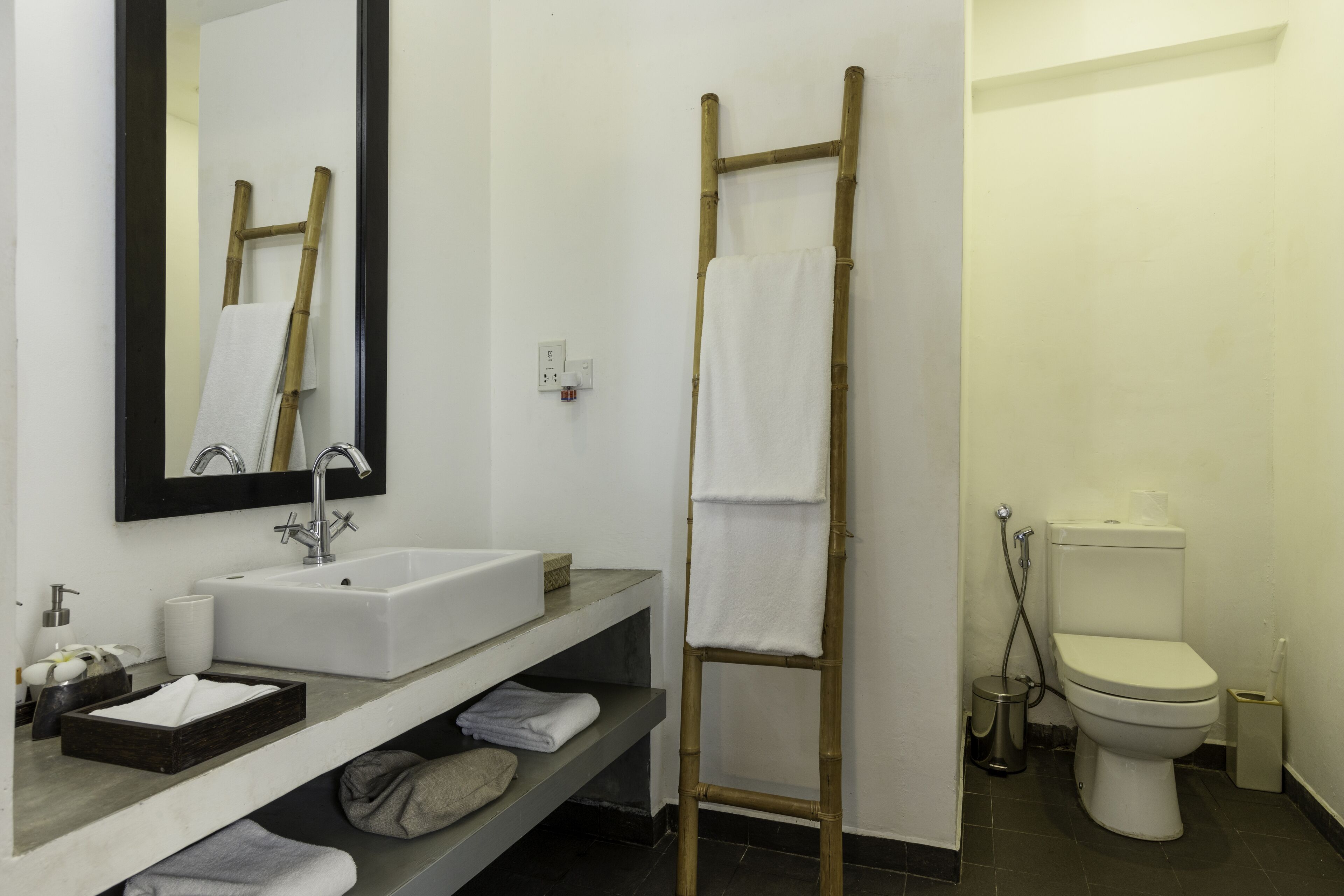 Een badkamer met een witte wastafel, spiegel en toilet. Er staat een houten ladder tegen de muur en een plank met handdoeken.