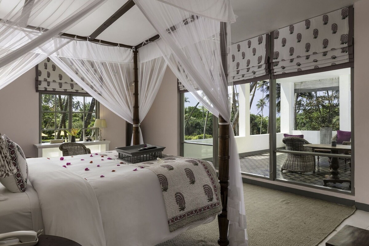 A bedroom with a four-poster bed, a large window with a view of palm trees, and a balcony with wicker furniture.