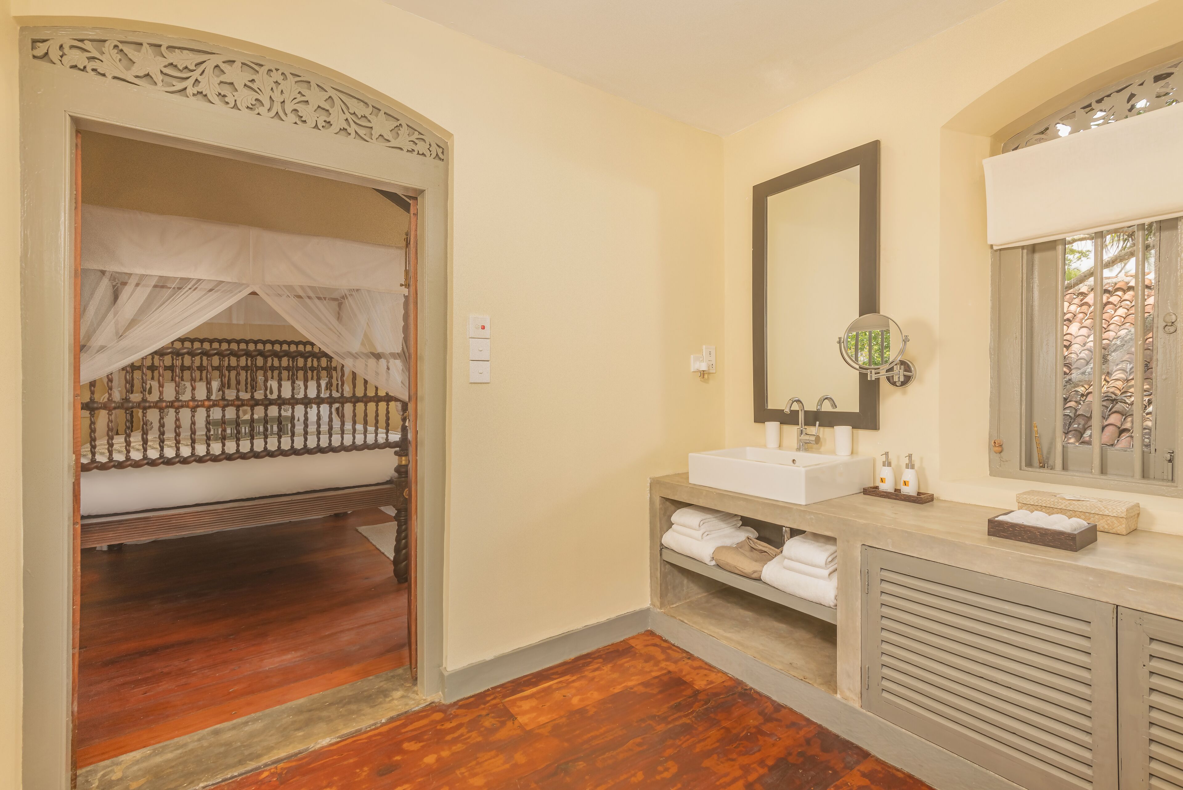 A room with a wooden floor, a bed with a canopy, a sink, and a mirror.