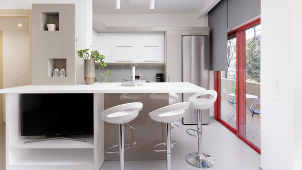A modern kitchen with white cabinetry, a central island with bar stools, and a large window with a red frame. -  A Luxury Retreat in Corfu Town by Konnect, Two-Bedroom Apartment (Corfu)