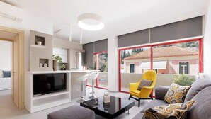 A modern living room with a flat-screen TV, a glass coffee table, and a sofa with patterned cushions. There is a yellow armchair and a dining area with a white table and chairs. -  A Luxury Retreat in Corfu Town by Konnect, Two-Bedroom Apartment (Corfu)