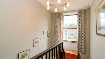 Uma escadaria com corrimão de madeira, um candelabro com quatro lâmpadas e uma janela com cortinas estampadas.