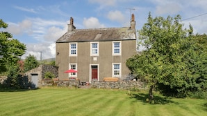 Une maison à deux étages avec une façade en pierre, une porte rouge et un petit jardin agrémenté d’une pelouse et d’un arbre.
