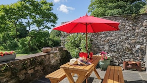 Une table de pique-nique en bois, surmontée d’un parasol rouge, entourée de murs en pierre et de plantes en pot.