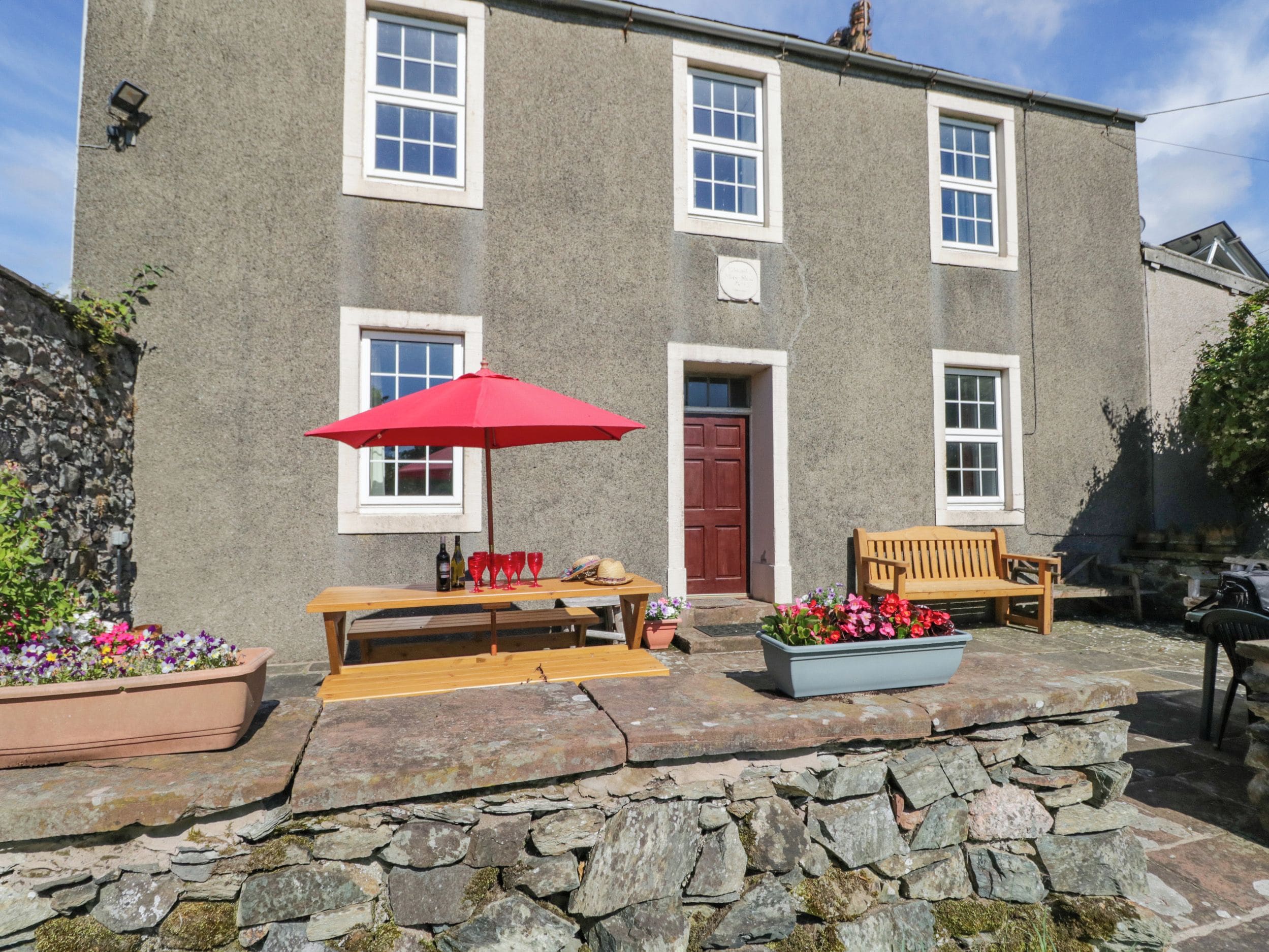 A stone building with a red door, a wooden bench, a red umbrella, and flower planters.