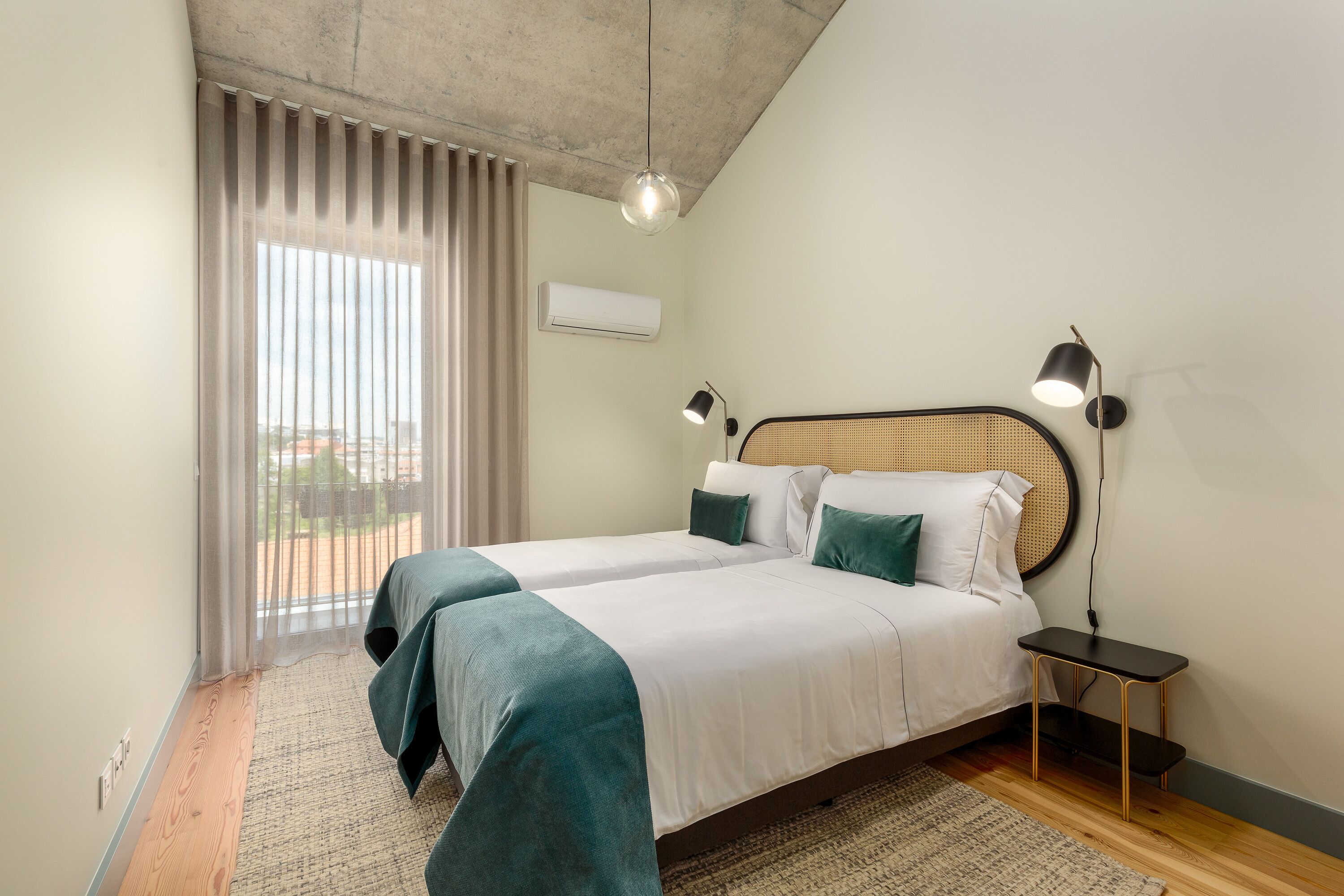 A hotel room with two beds, a large window with curtains, an air conditioning unit, and a small table with a lamp.