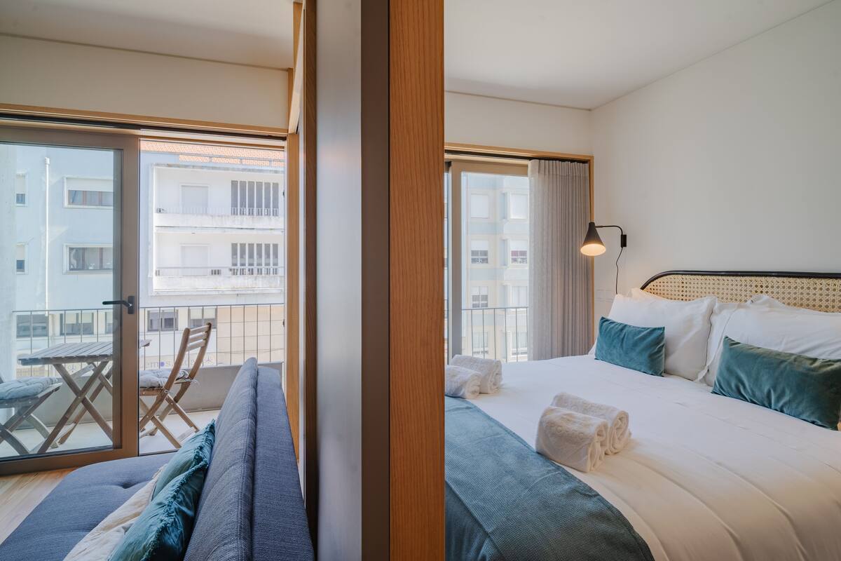 Une chambre d’hôtel avec un lit, un canapé, un balcon avec une table et des chaises, et une vue sur les bâtiments.