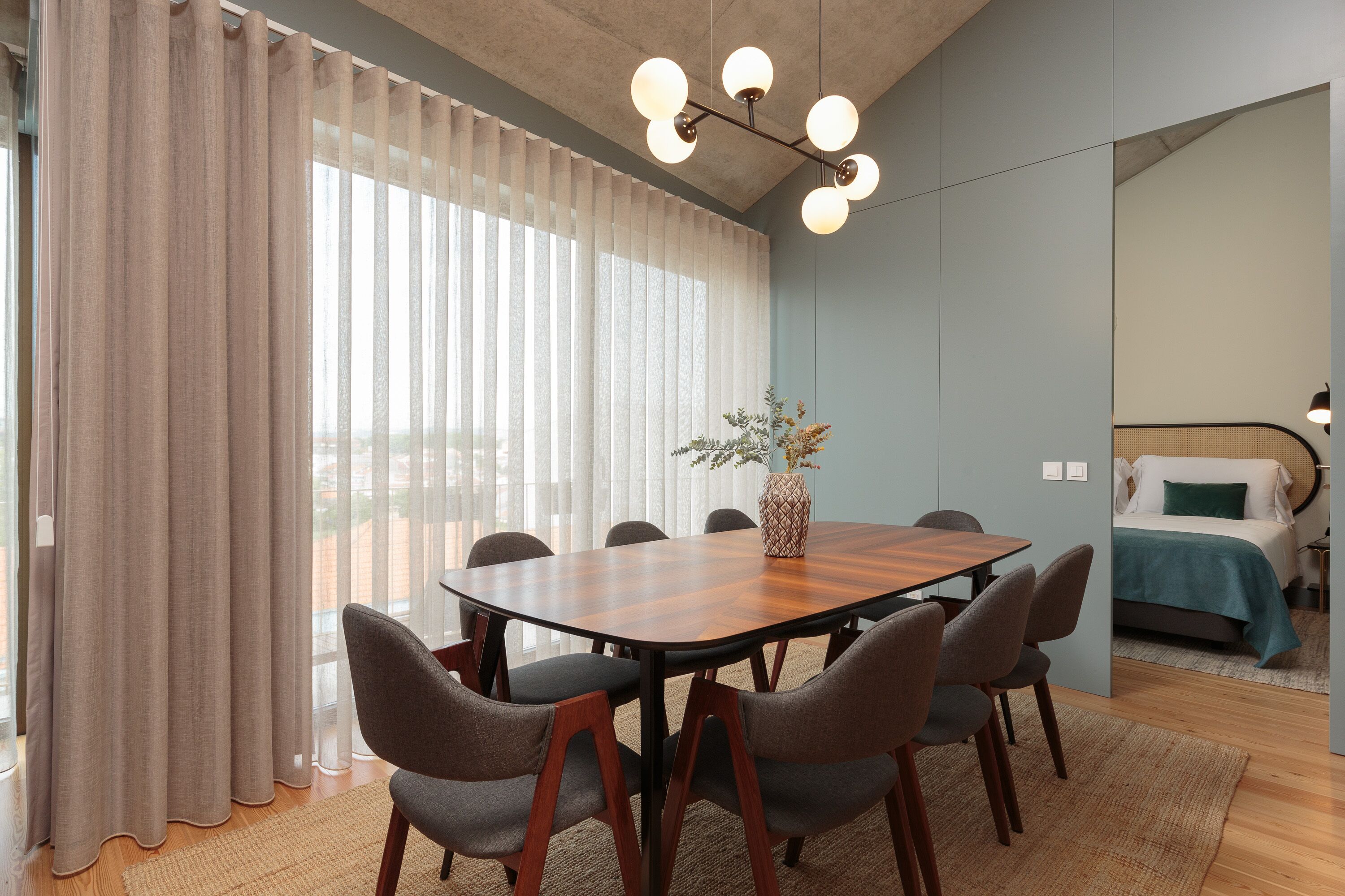 Una habitación moderna con una mesa de madera, seis sillas, un jarrón con flores y una cama con almohada verde.