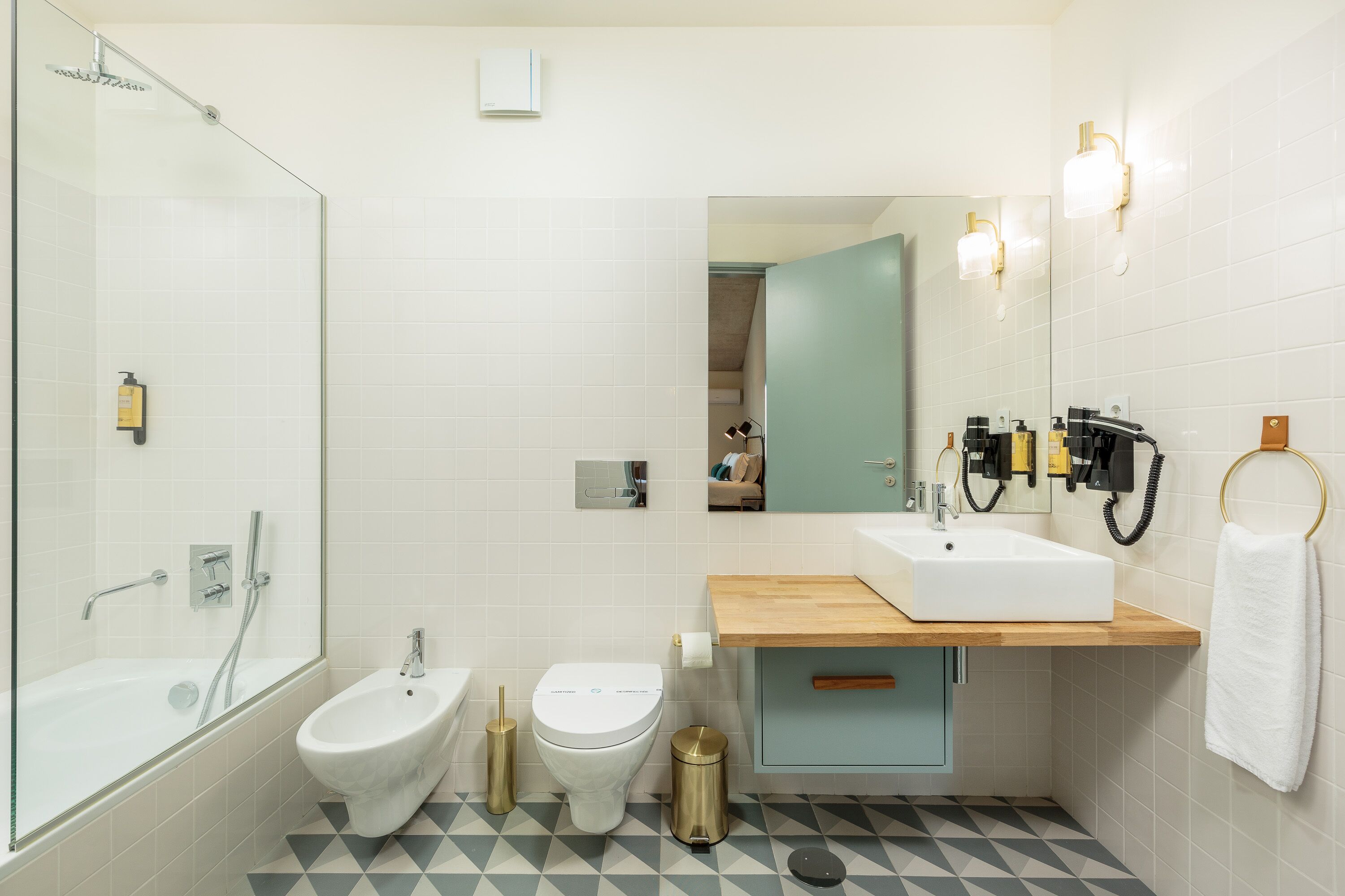 A modern bathroom with a glass shower enclosure, a bidet, a toilet, a sink with a wooden countertop, and a mirror with wall-mounted lights.