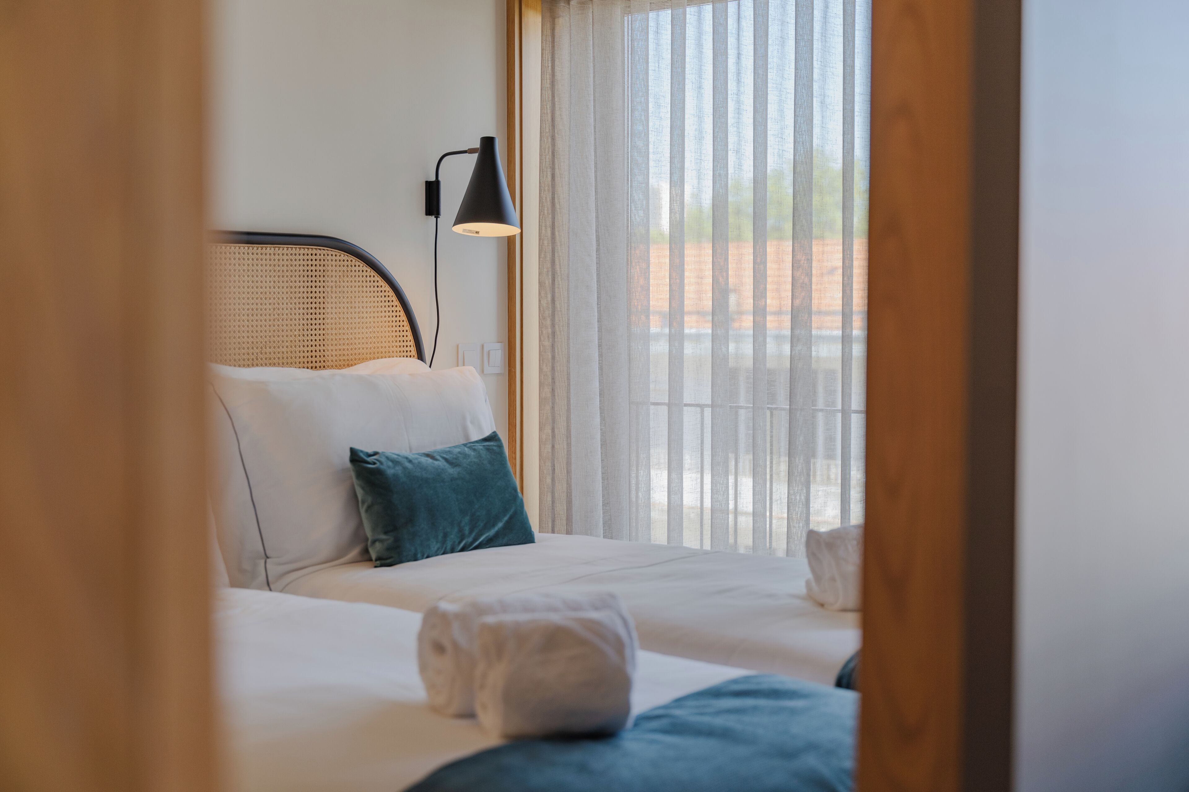 A neatly made bed with white linens, a teal pillow, and a rolled towel. A black desk lamp is on the bedside table.