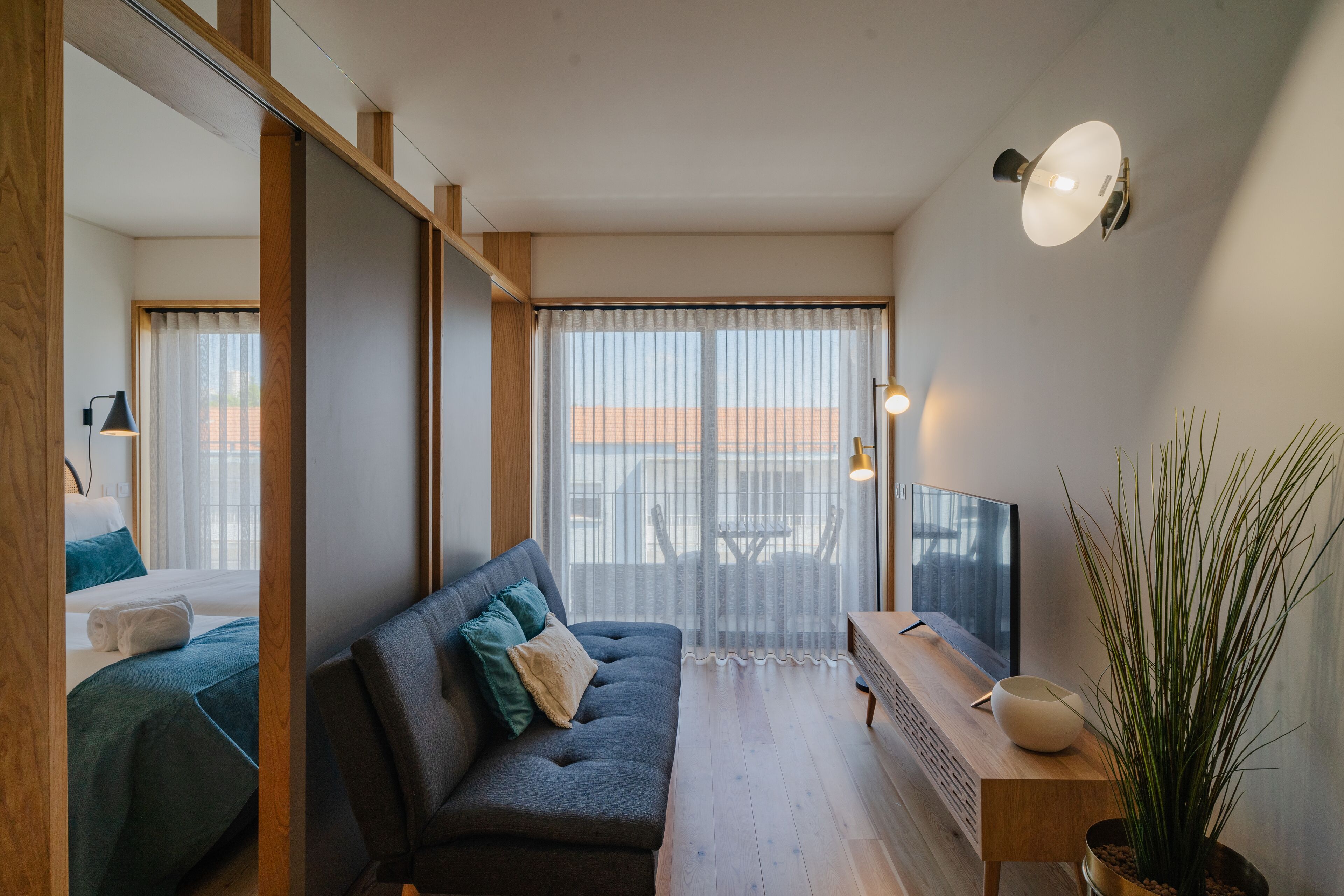 A modern living room with a sofa, a TV, a wooden side table, and a large window with sheer curtains.