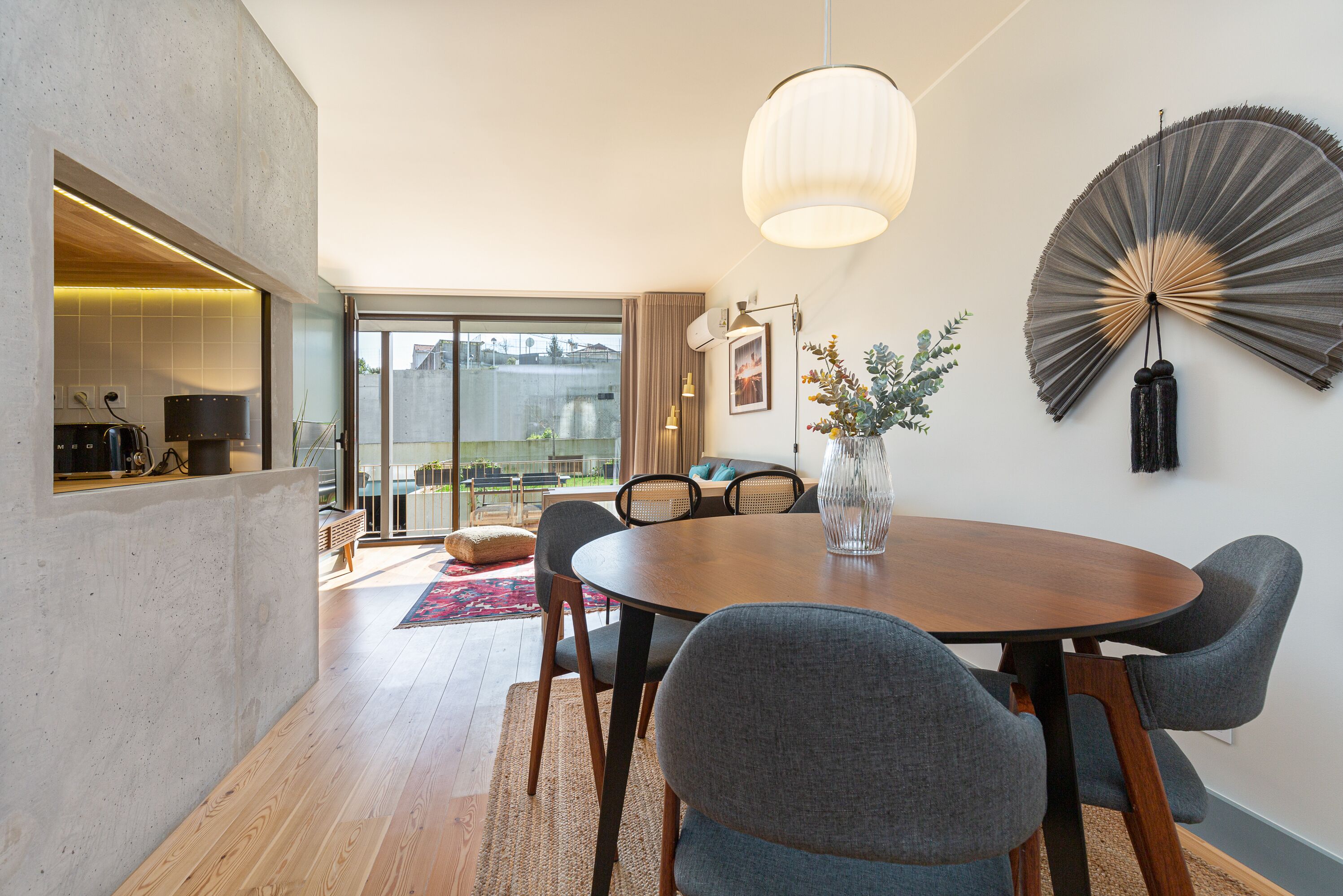 A modern dining area with a round wooden table, grey upholstered chairs, a vase with flowers, and a large wall fan.