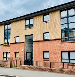 A multi-story brick building with large windows and a black metal fence in front. - Modern 2 Bedroom Apartment With Free Parking (Glasgow)
