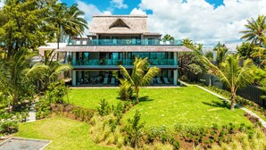 Un bâtiment moderne à deux étages, doté d’un toit de chaume, entouré d’une végétation luxuriante et de palmiers.