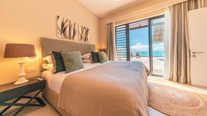 Une chambre d’hôtel avec un lit, une table de chevet, une lampe, un tableau au mur et une vue sur la plage par la fenêtre.