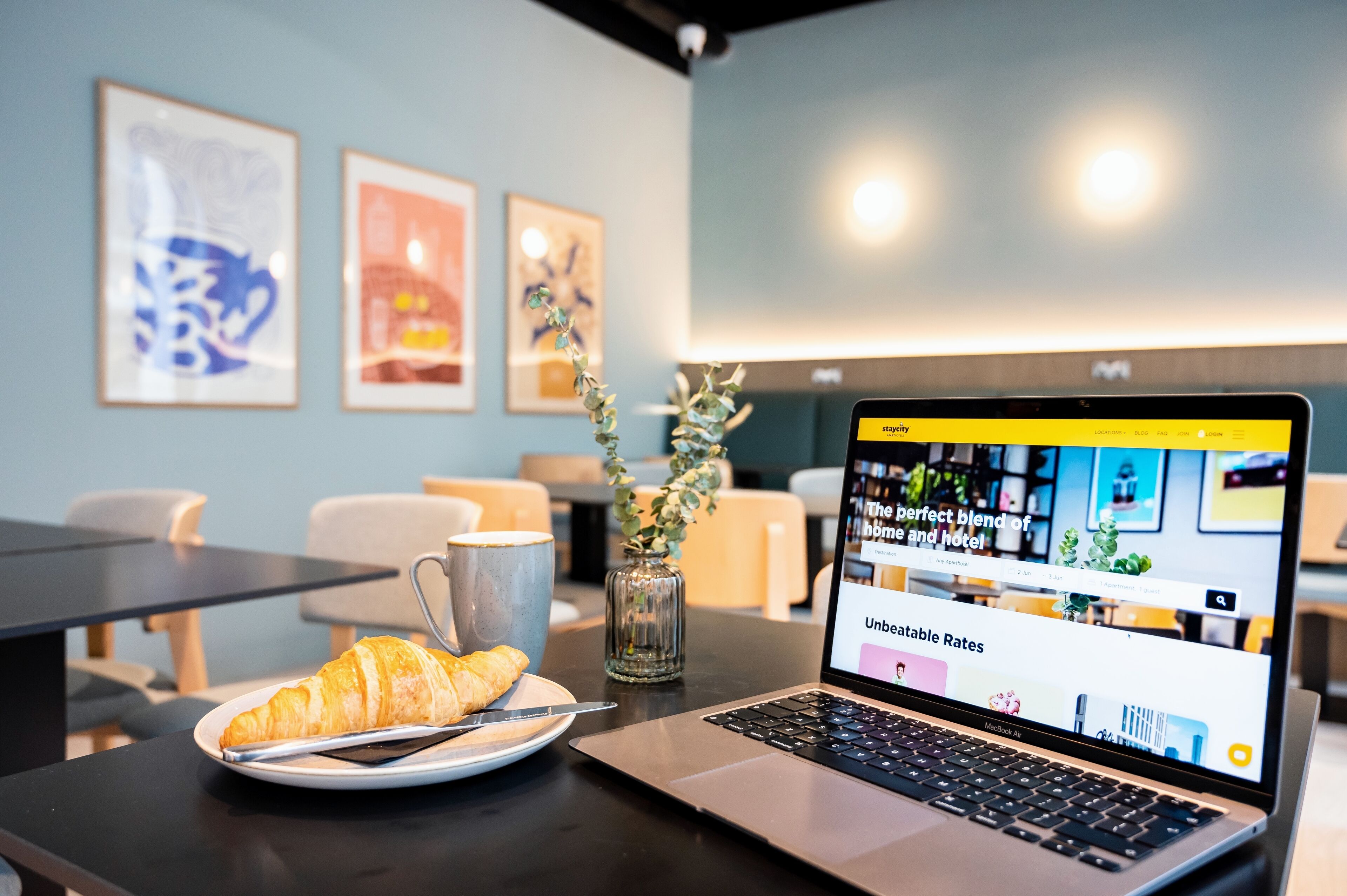 A laptop on a table with a croissant and a mug, framed artwork on the wall, and a vase with greenery.