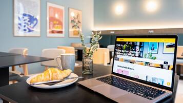 A laptop on a table with a croissant and a mug, framed artwork on the wall, and a vase with greenery.