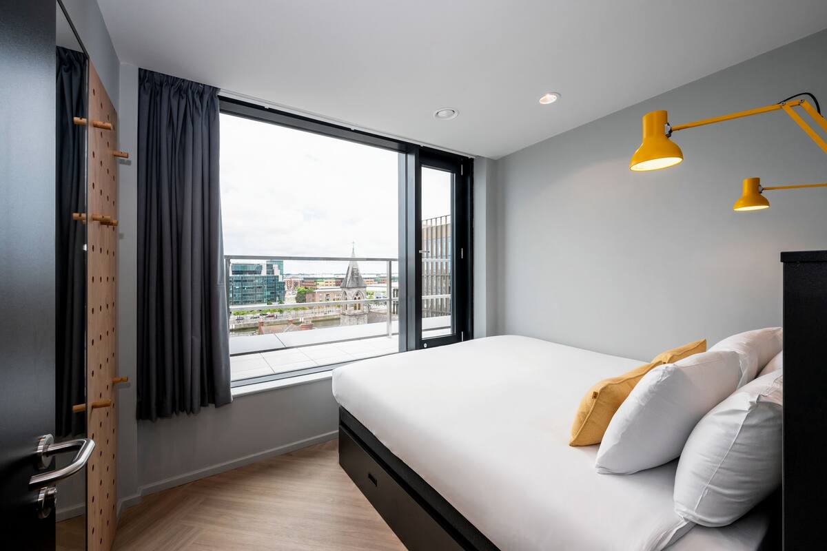 A modern bedroom with a large window, a bed with white and yellow pillows, and two yellow bedside lamps.