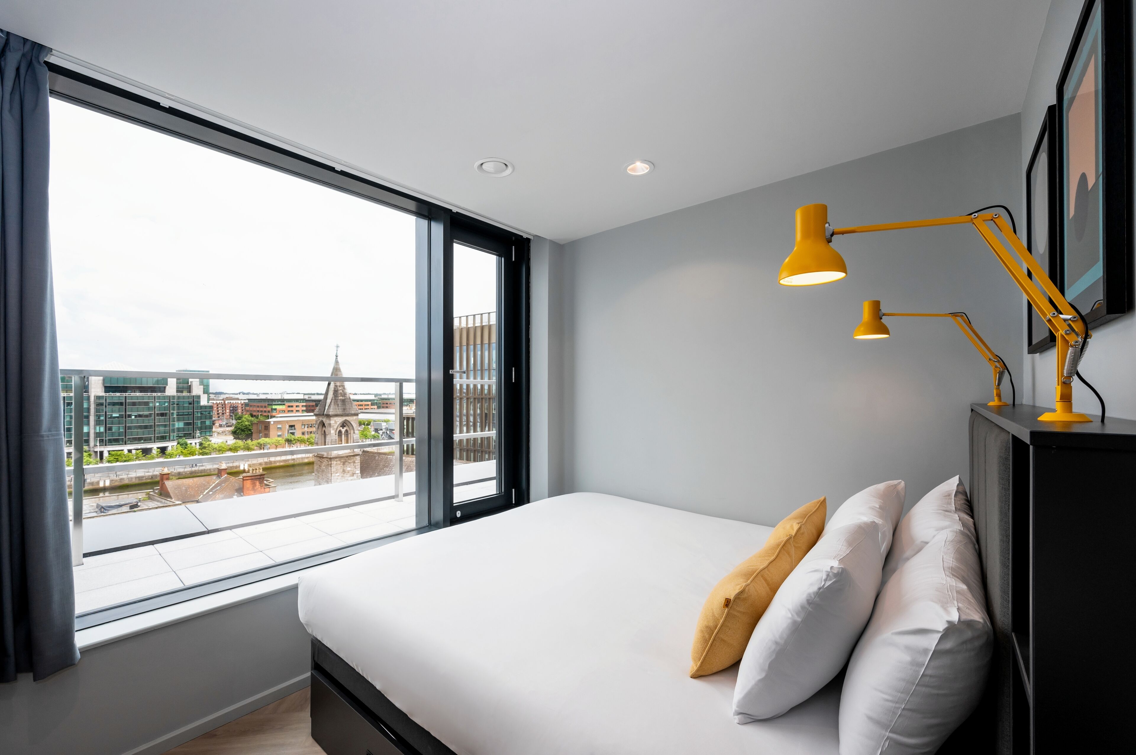 A modern hotel room with a large bed, a desk with a yellow lamp, and a view of the cityscape through a large window.