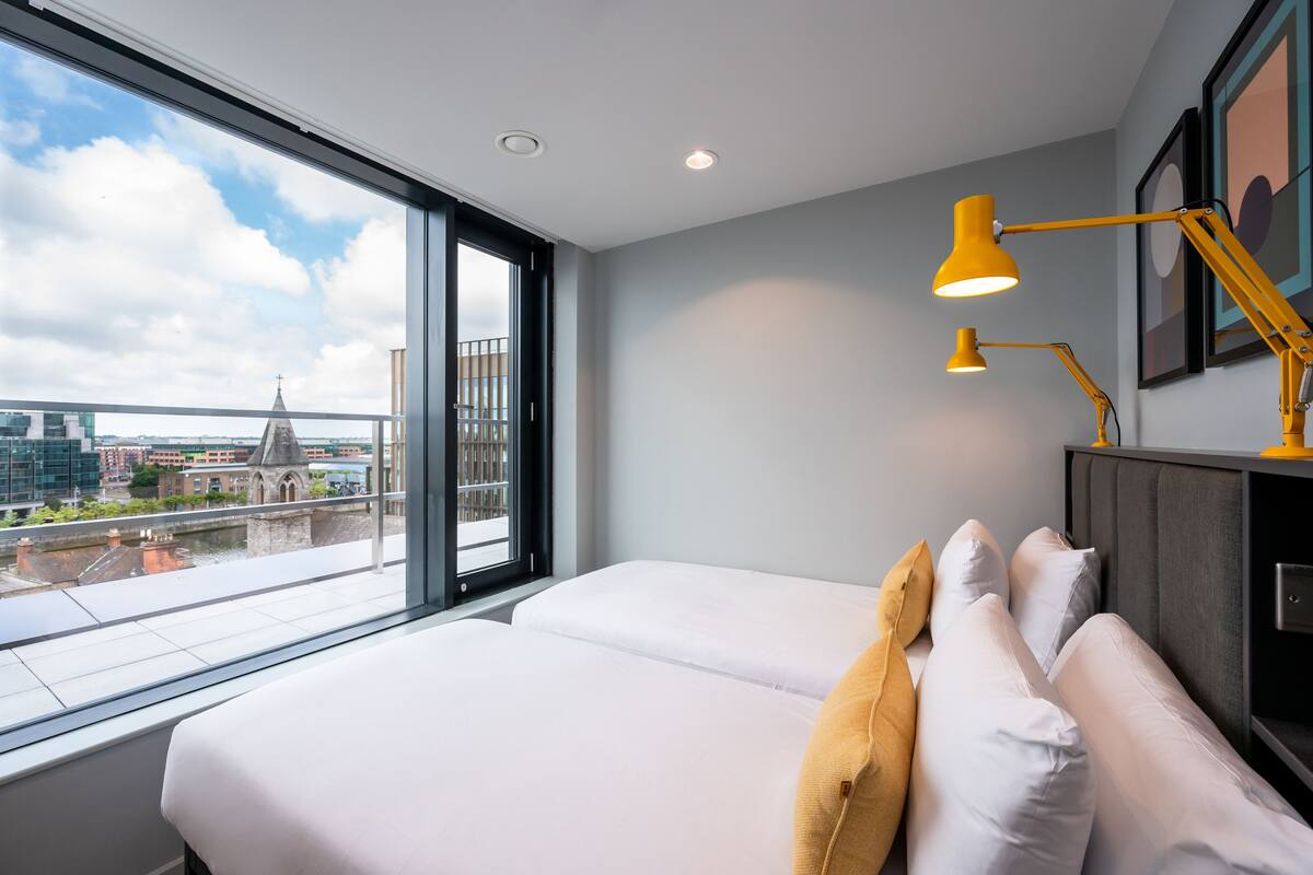 A modern hotel room with a large window, two beds, a desk, and a yellow desk lamp.