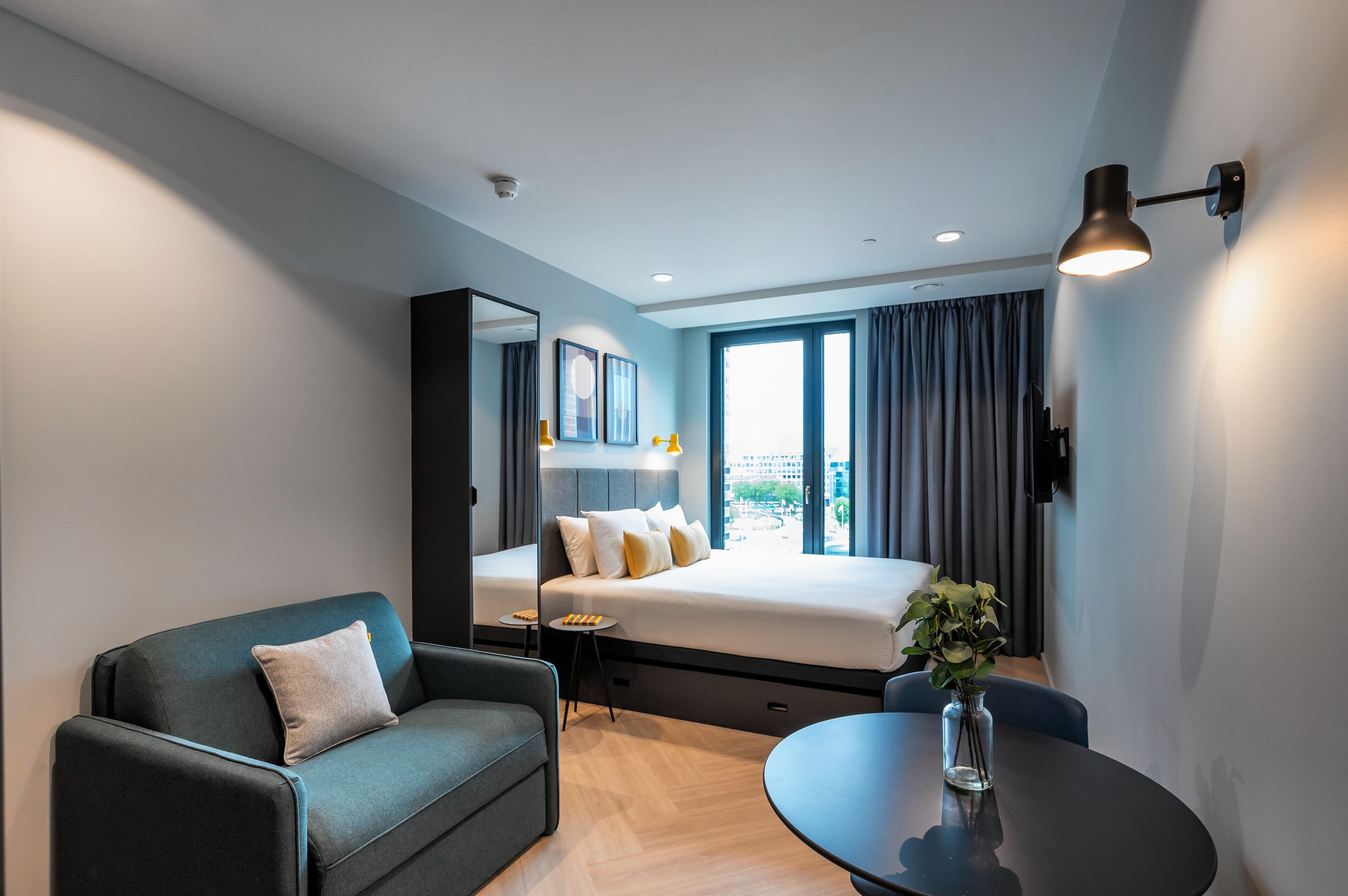 A modern hotel room with a bed, a sofa, a small table, and a large window with curtains.