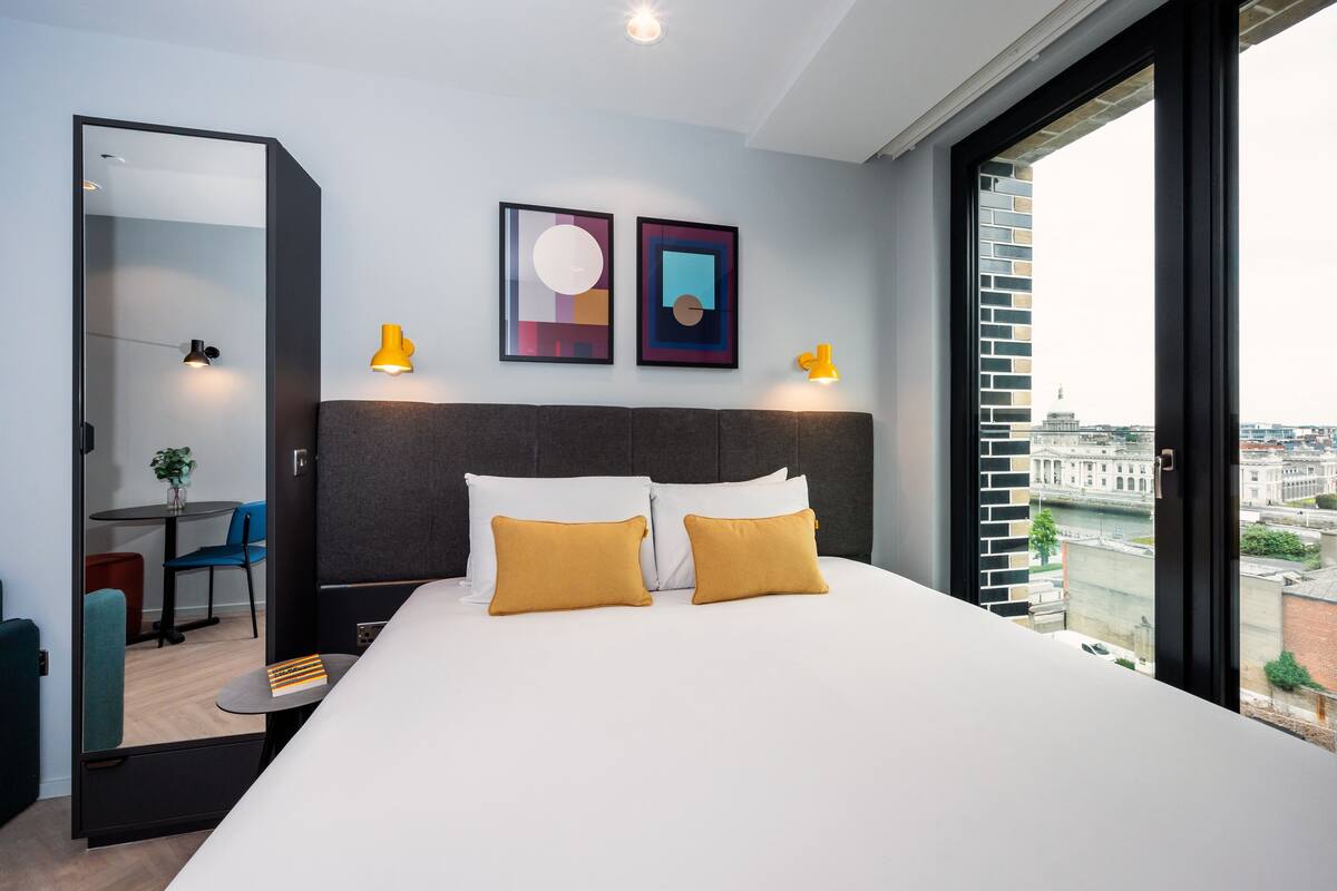 A modern hotel room with a large bed, two pillows, a headboard, and a city view.