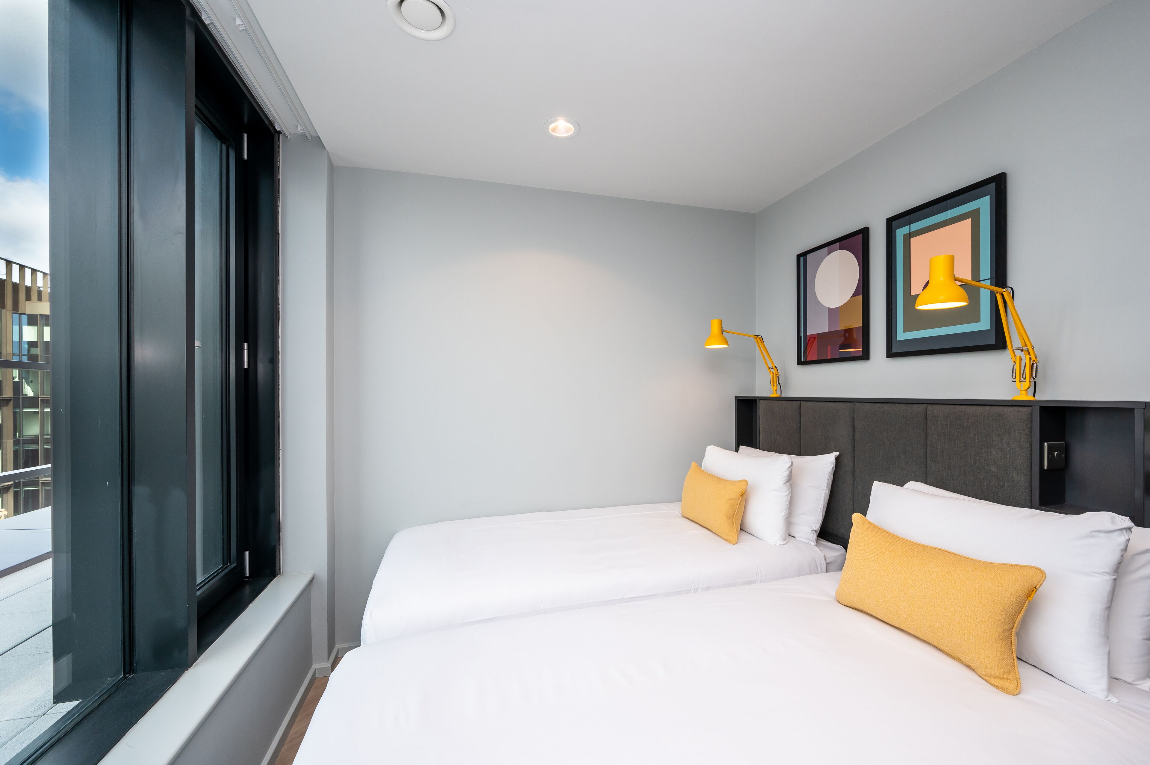 Une chambre d’hôtel avec deux lits, une grande fenêtre et deux lampes fixées au mur.