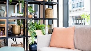 Een moderne zithoek met een grijze bank, een boekenplank vol boeken en decoratieve objecten, en een groot raam met uitzicht op een gebouw.