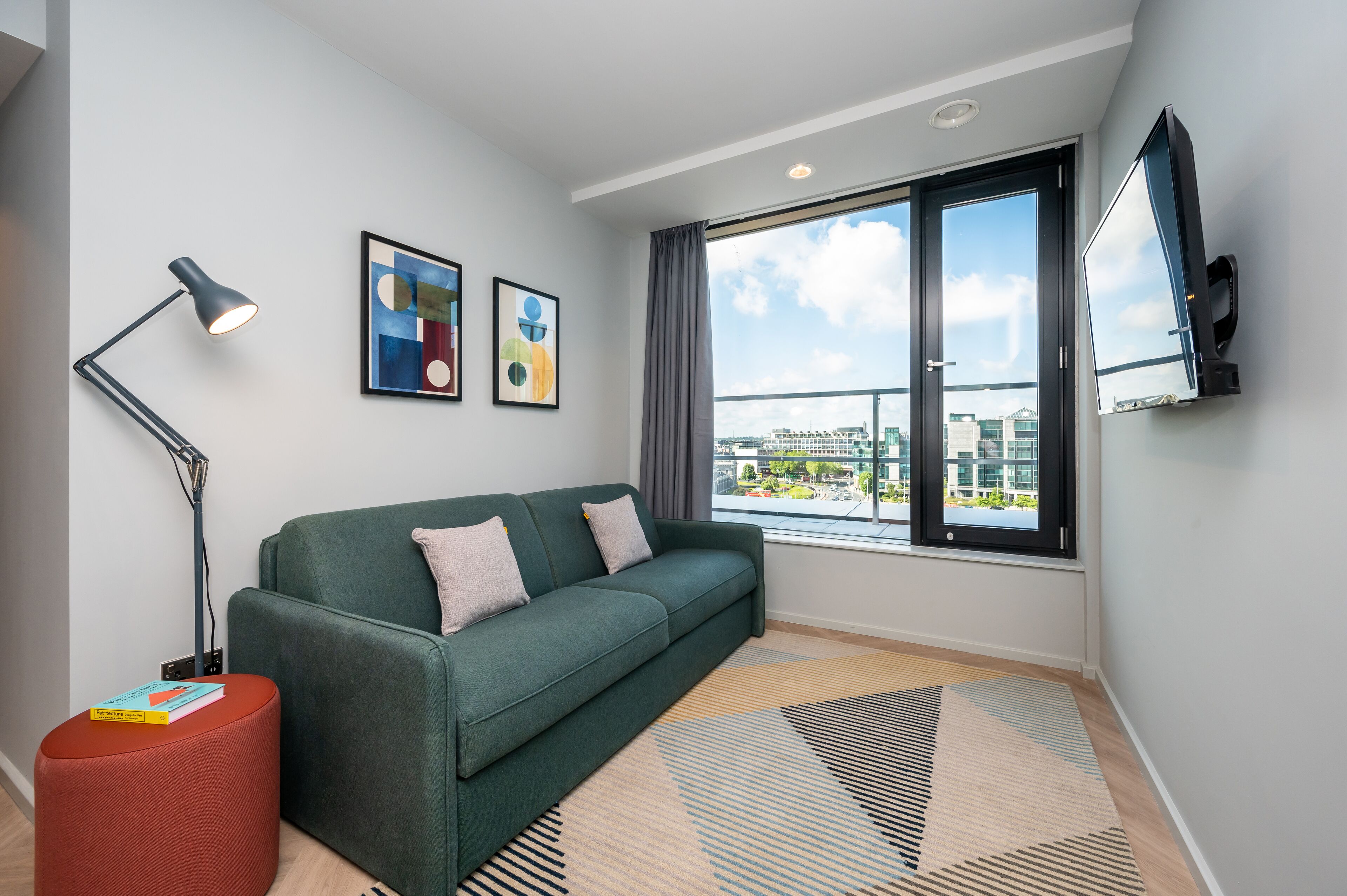 A modern living room with a green sofa, a red ottoman, a floor lamp, and a large window with a city view.