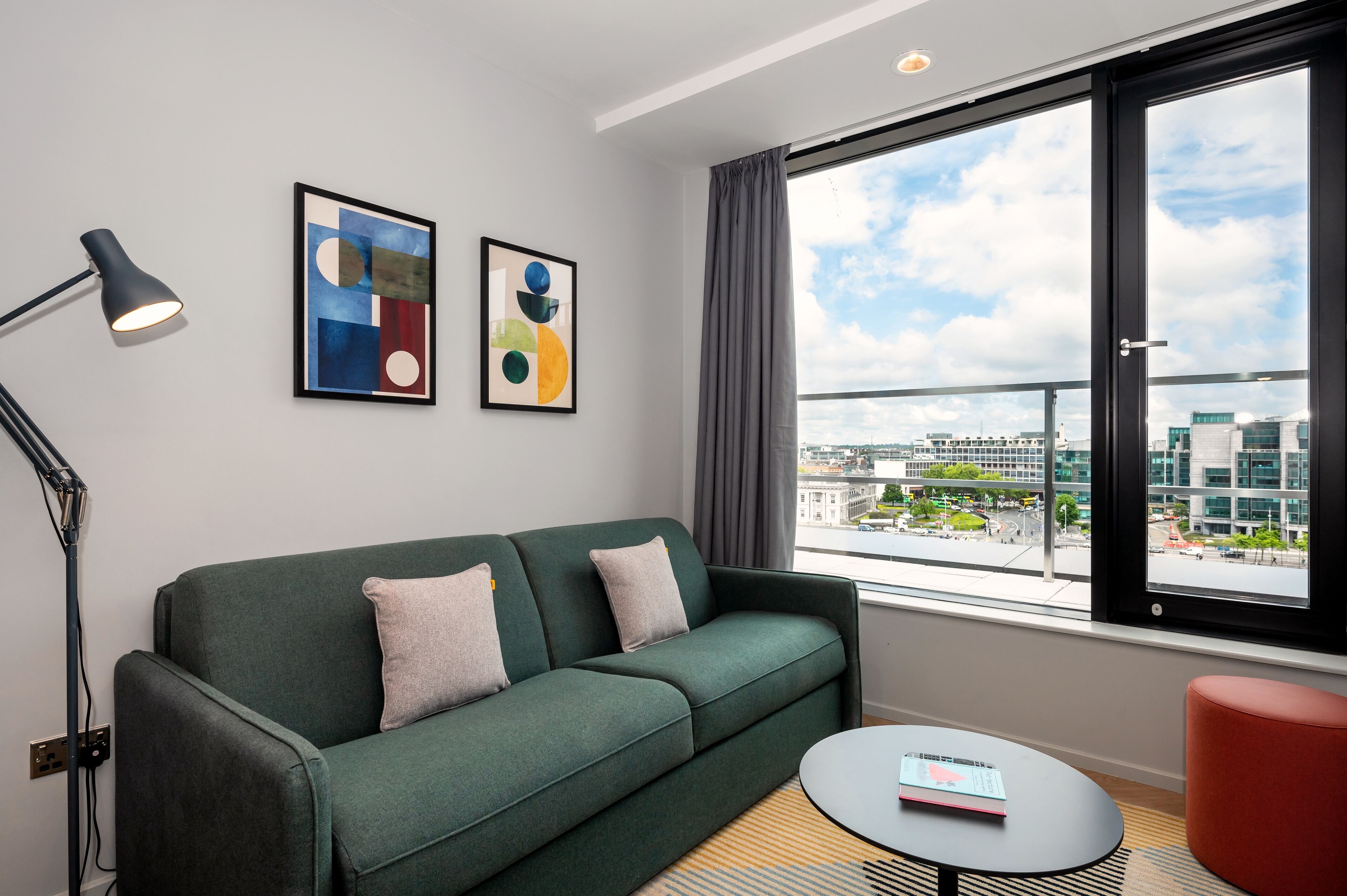 A modern living room with a green sofa, a small round table, and a view of a cityscape through large windows.