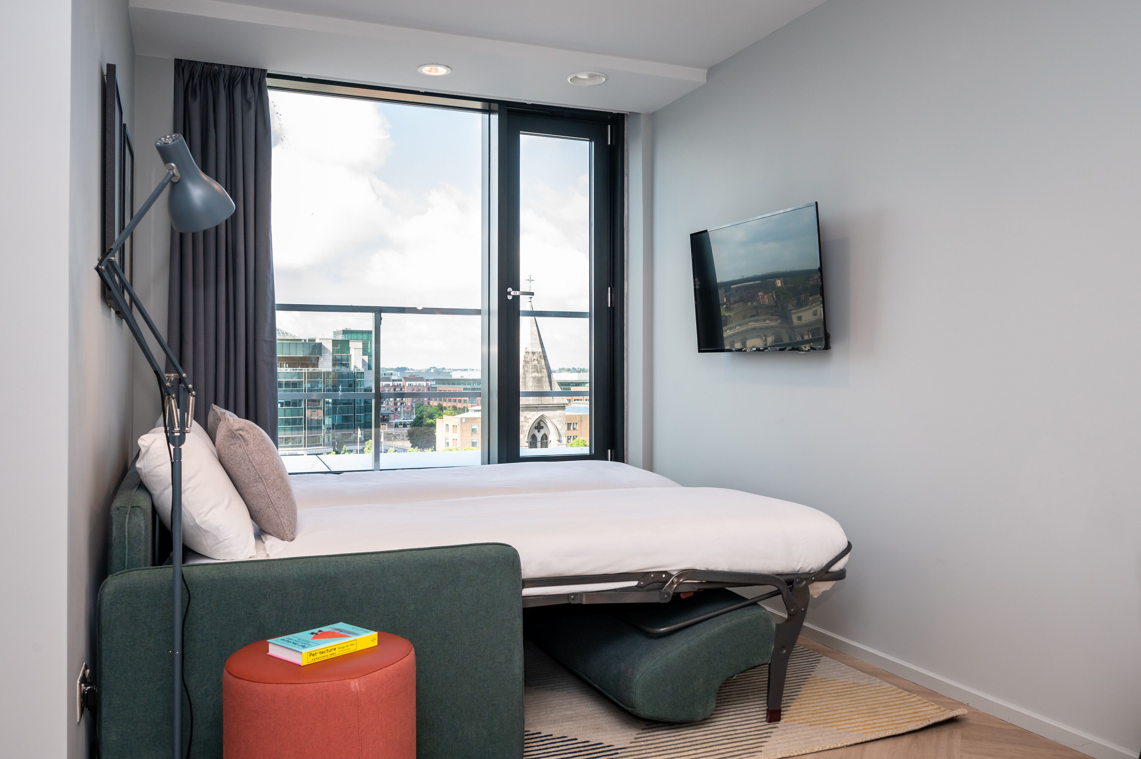 A modern hotel room with a bed, a desk, a chair, a TV, and a large window offering a city view.