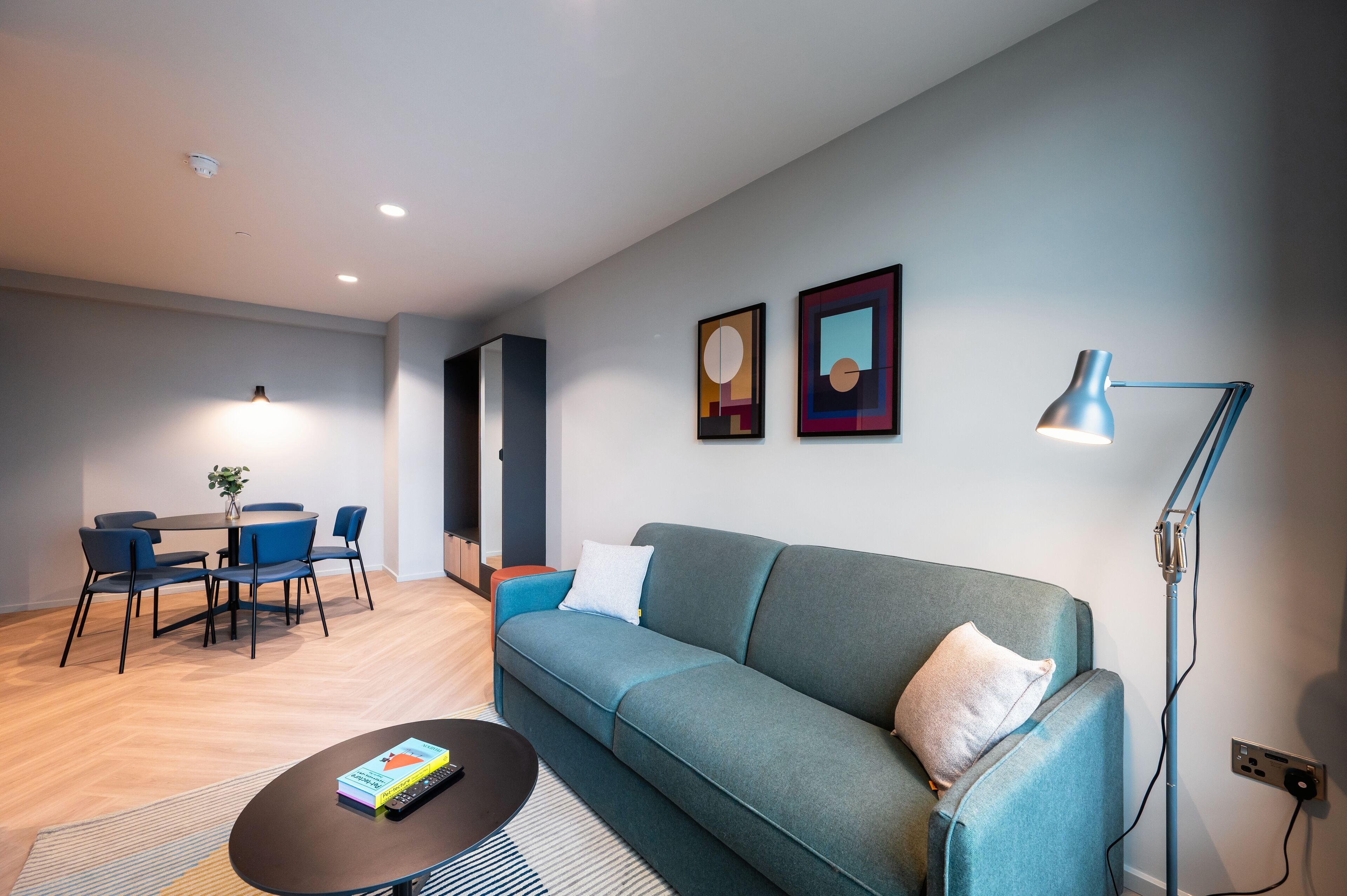 A modern living room with a teal sofa, a round coffee table, a dining area with a table and chairs, and a blue floor lamp.